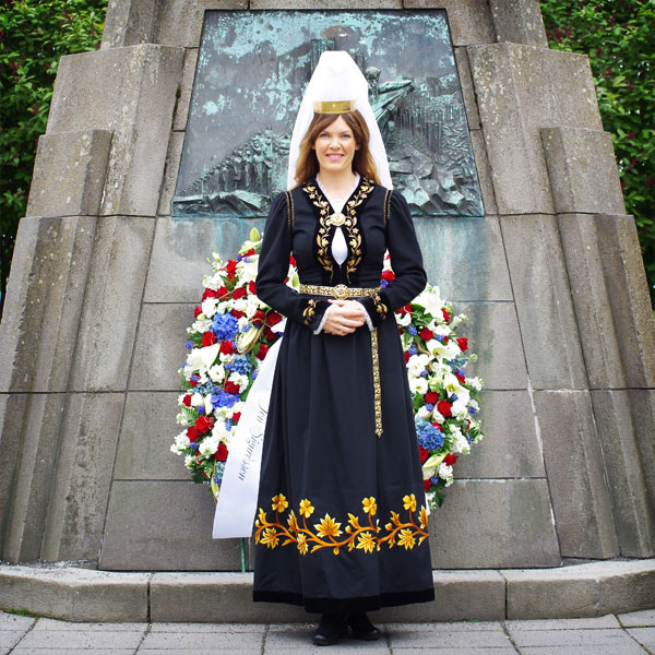The Mountain Lady stands in front of a podium decorated with a garland of flowers. Her dress is black, embroidered with golden flowers, she is wearing a high white headdress and a tiara.