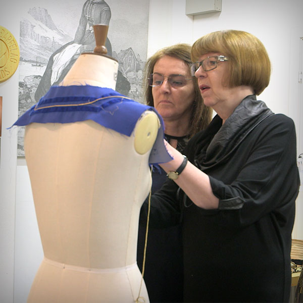 Two women looking closely at a piece of sewing they are doing on a mannequin.
