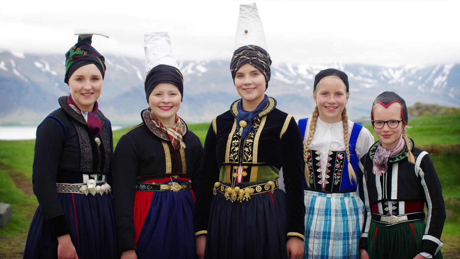 A group of three young women and two girls, all dressed in multicolored costumes of the older style. In the background a green slope and a mountain range.