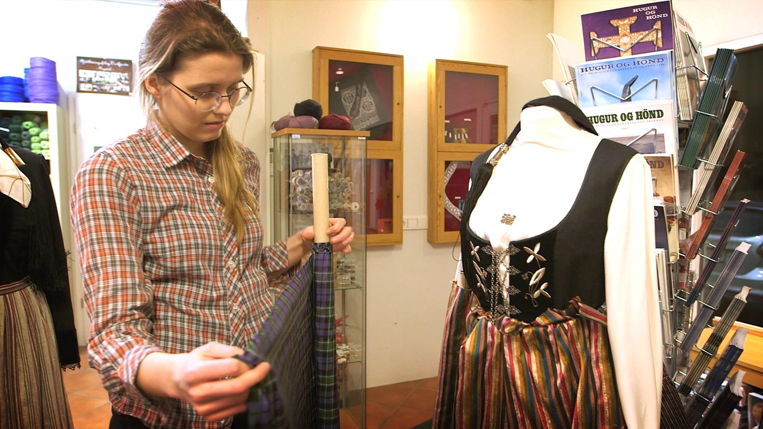 A young woman stands in front of a mannequin dressed in 20th century national dress. She is choosing between two kind of cloth for an apron.