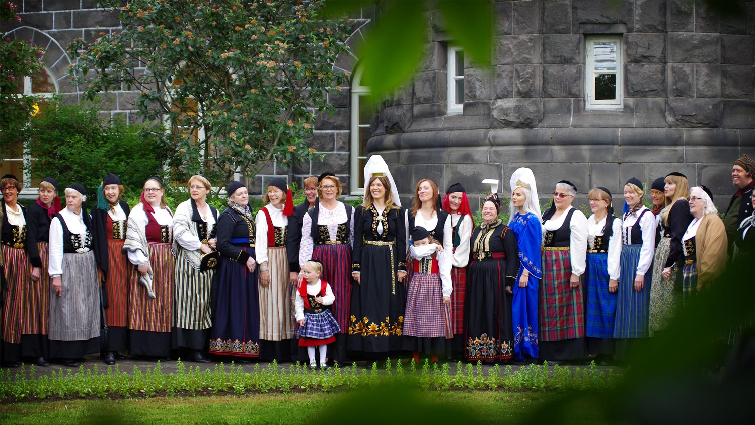 A group of many women in a long line, dressed in all kinds of national costume,  in the garden behind the Icelandic parliament, Althingi. In the middle is the Fjallkonan, the Icelandic Mountain Woman, a young woman dressed as a symbol for the country.