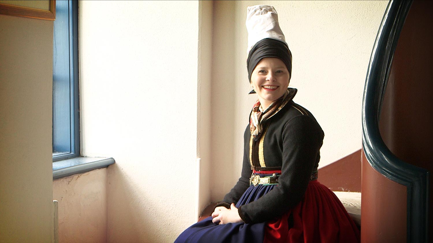 A young woman sitting on stairs in front of a window smiling in the direction of the camera. She is dressed in a red skirt, blue apron, and dark jacket and wears a high, white headdress.