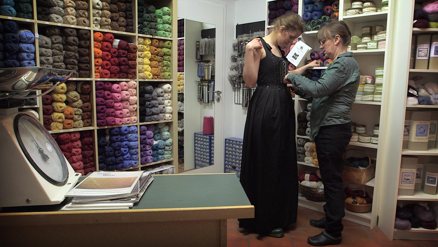 A young woman trying on the dress she is sewing with the help of an older woman, her coach. They are surrounded by shelves stacked with yarn of many colors.