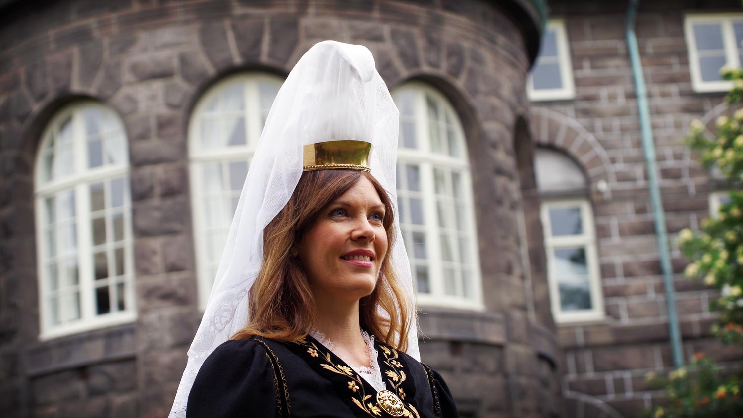 A portrait of a young woman wearing a dress with high headdress and tiara; a veil falling to her shoulders. She is the Mountain Woman on the National Day, Mountain Woman being the symbol for Iceland. In the background is the parliament house.
